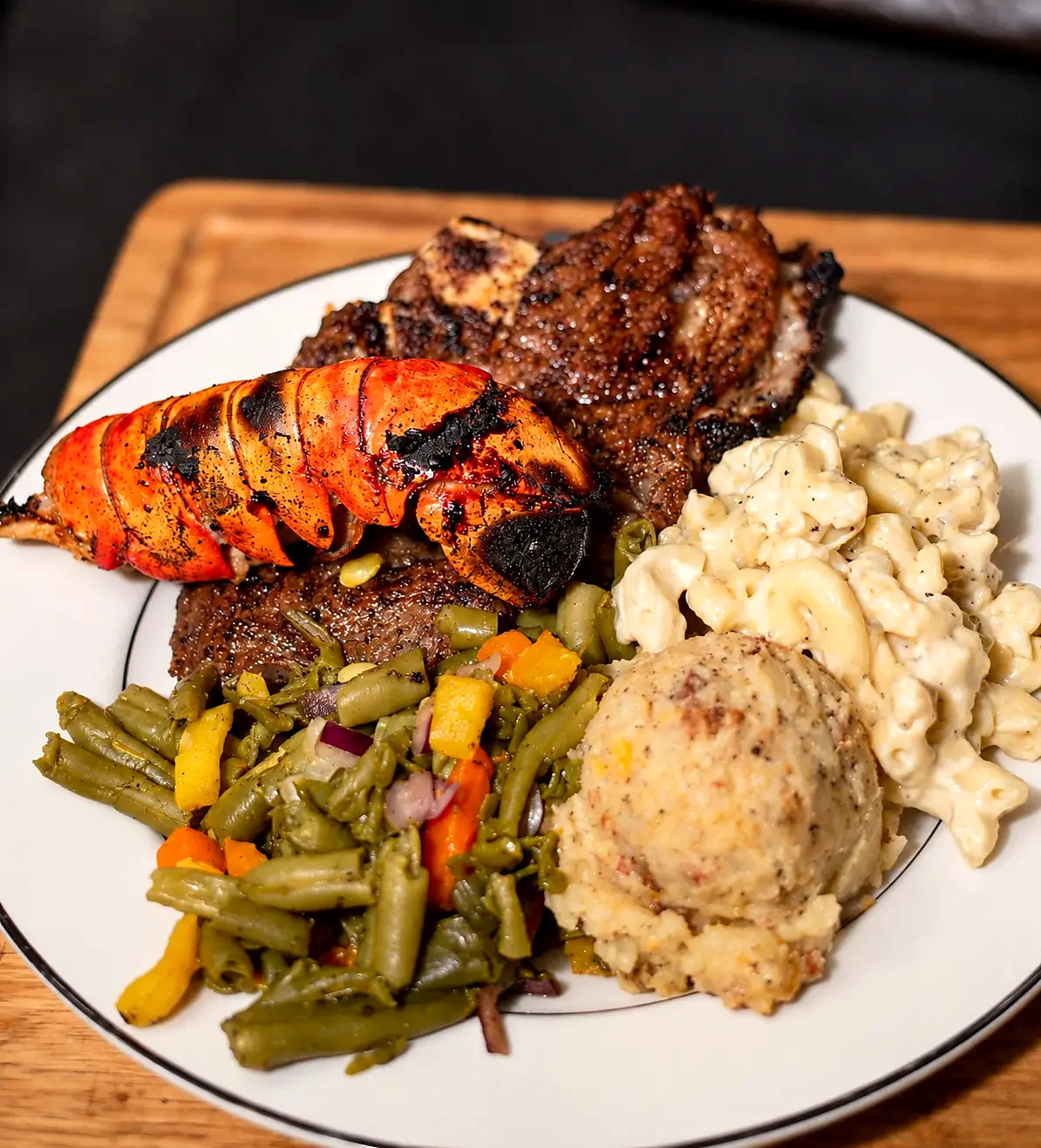 A catered surf and turf plate: char-grilled lobster tail over a bone-in steak with a vegetable medley, loaded mashed potatoes and mac and cheese