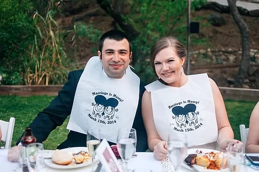 Bride and groom smiling over dinner in matching wedding bibs at an outdoor table