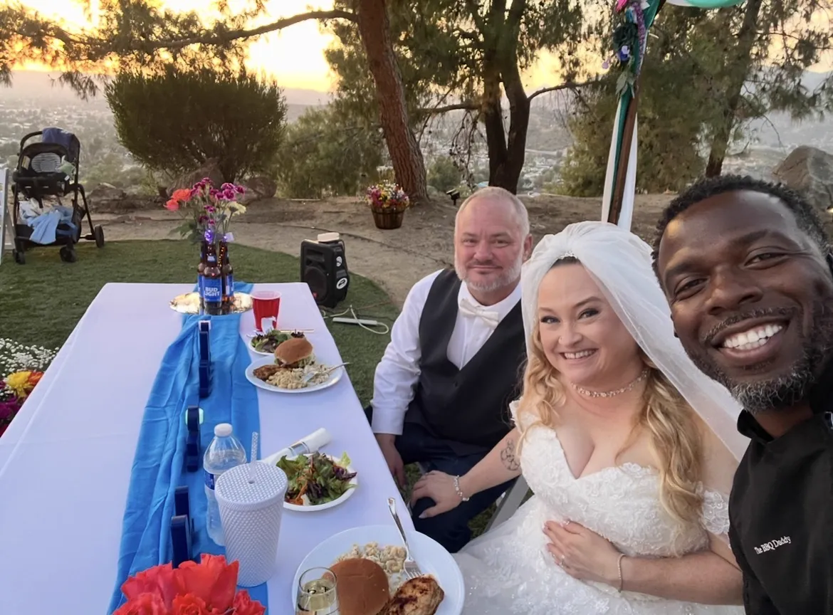 Newlyweds and Chef Terry at an outdoor reception table with dinner plates and flowers