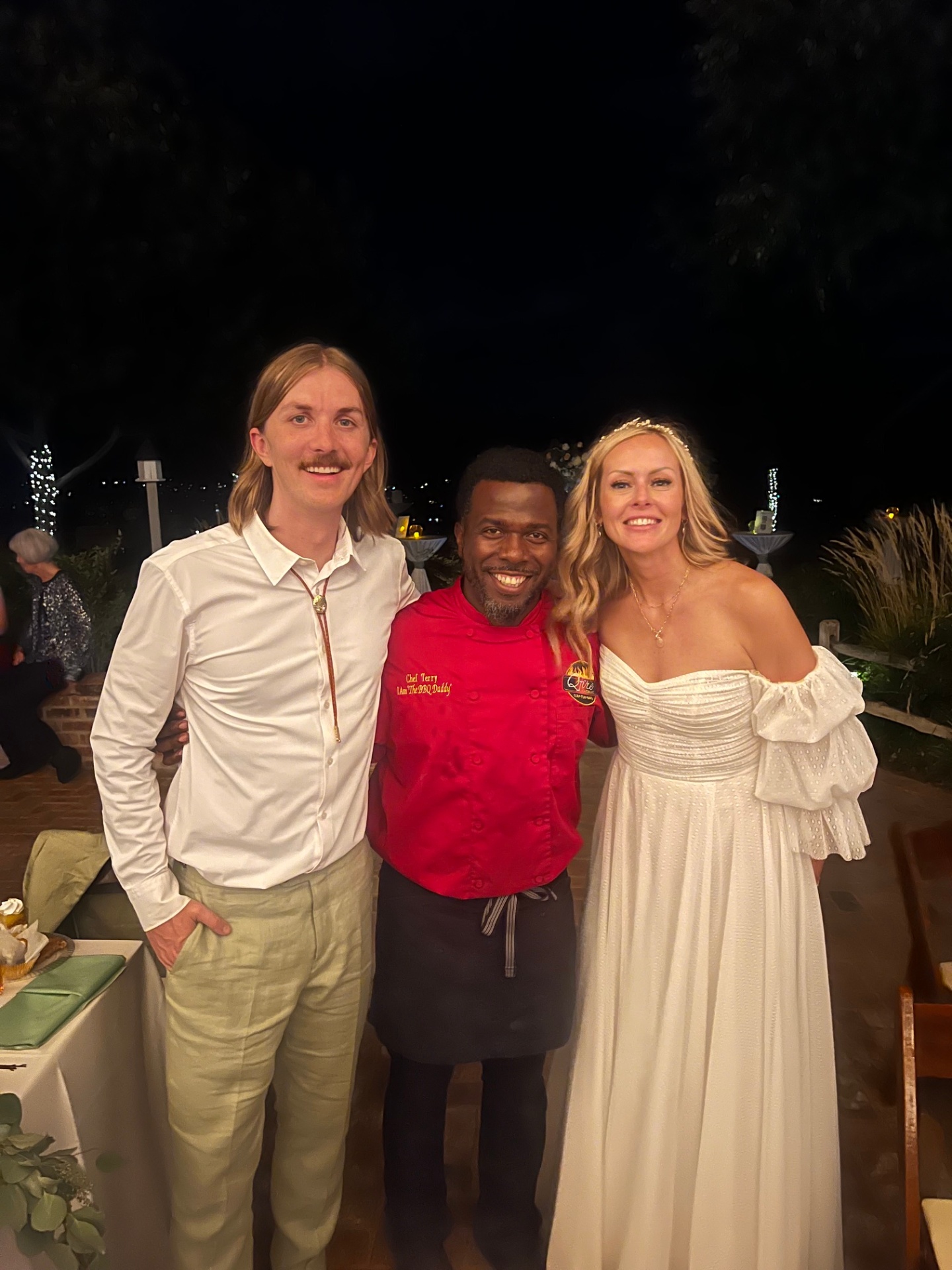 Chef Terry in red BBQ Daddy chef coat with bride and groom in bolo tie at an evening Phoenix Metro wedding reception