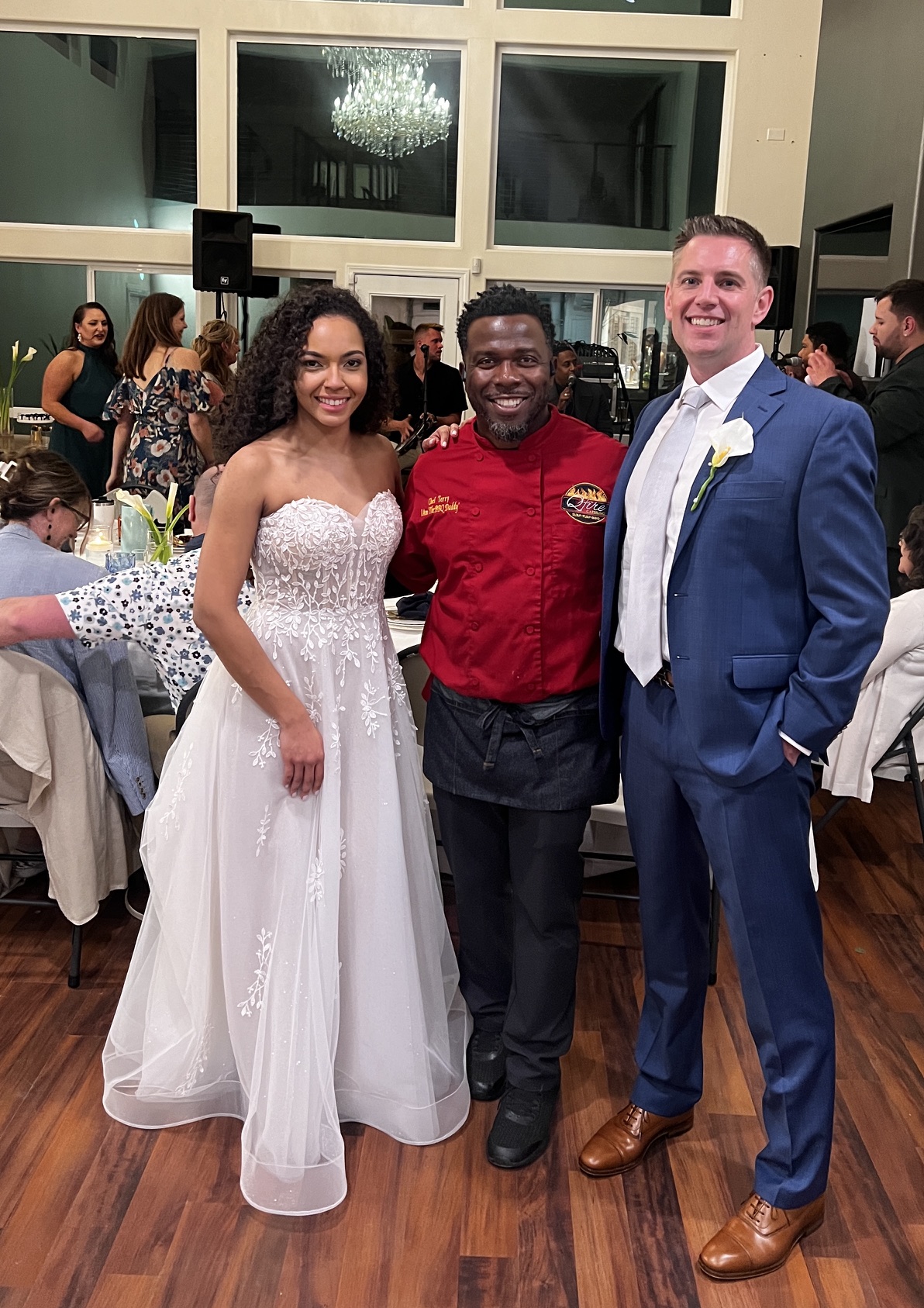 Chef Terry in red BBQ Daddy chef coat with bride and groom in blue suit at an indoor Phoenix Metro wedding reception