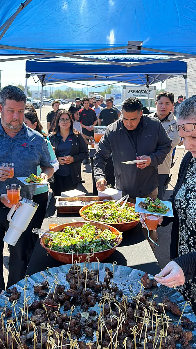 Employees lining up under a pop-up tent for grilled steak bites and fresh salads at an outdoor company lunch