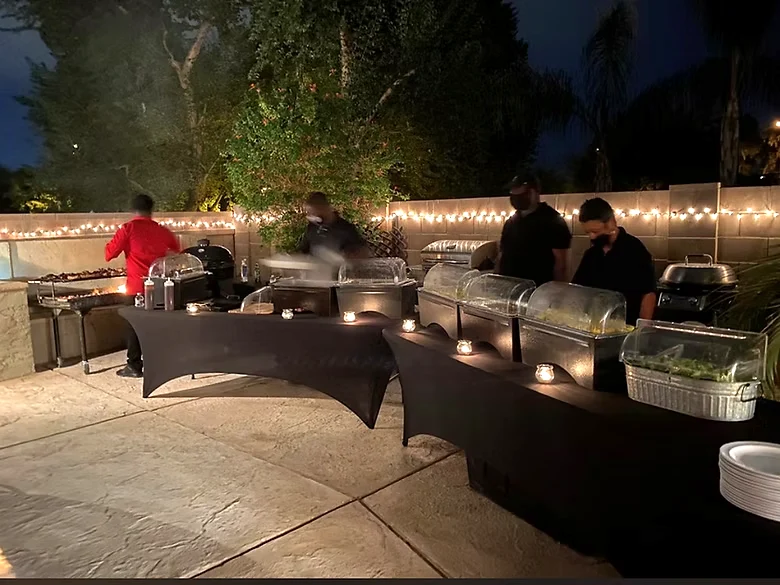 Catering team behind a patio buffet with covered chafing dishes and string lights at night