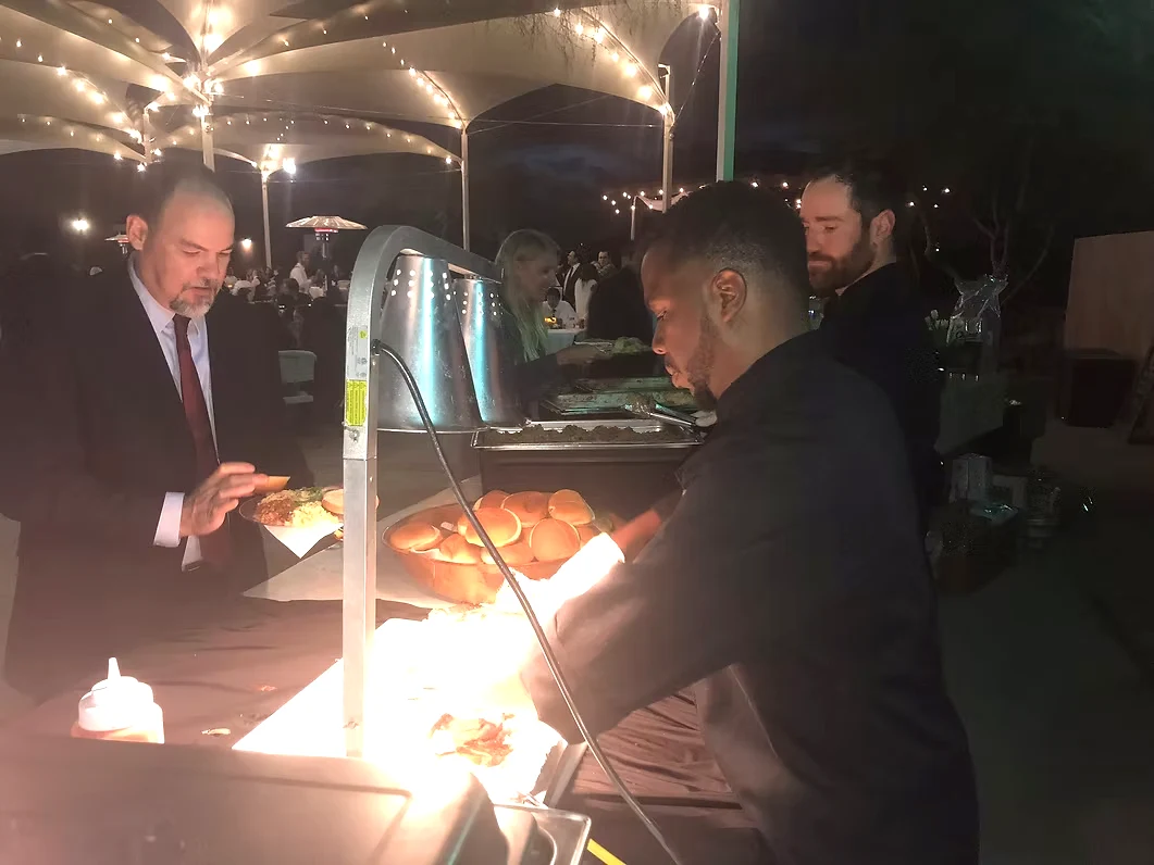 Catering attendant serving a wedding guest at a buffet beneath warm string lights