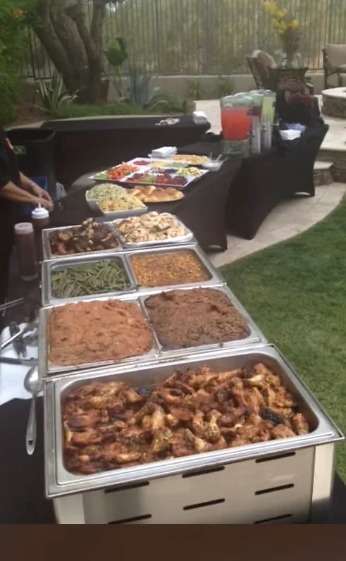 Outdoor buffet with trays of meats and sides, salad, rolls and colorful drink dispensers